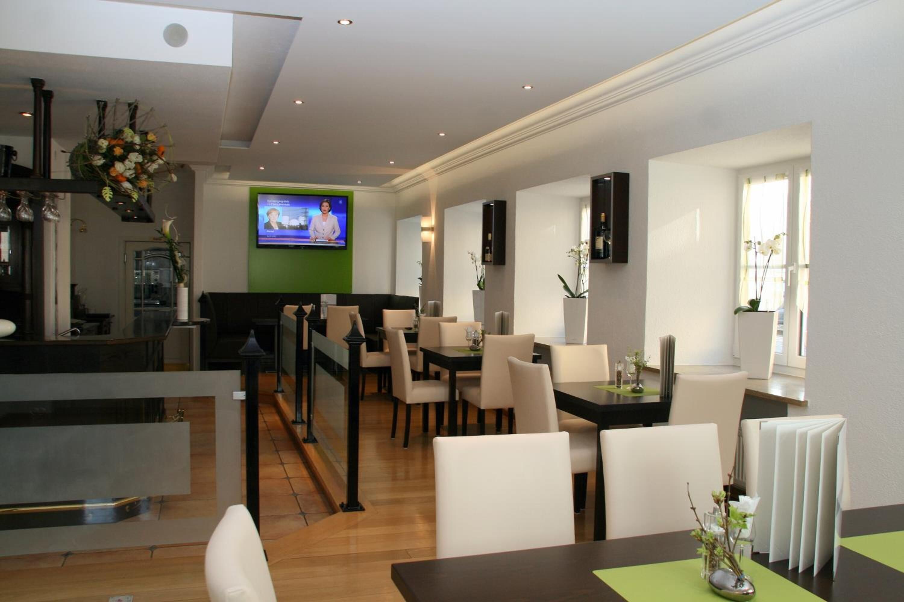 A modern hotel lobby with a reception desk, dining area with chairs and tables, a television screen, and decorative plants.