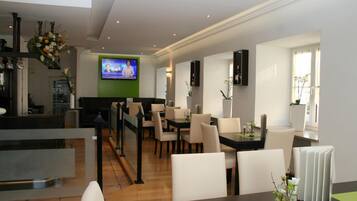 A modern hotel lobby with a reception desk, dining area with chairs and tables, a television screen, and decorative plants.