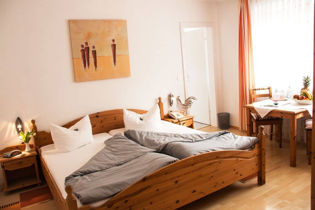 A neatly arranged bedroom with a wooden bed, a bedside table with a lamp, a small table with a fruit bowl, and a painting on the wall.