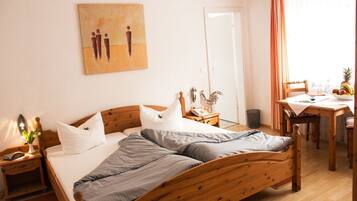 A neatly arranged bedroom with a wooden bed, a bedside table with a lamp, a small table with a fruit bowl, and a painting on the wall.