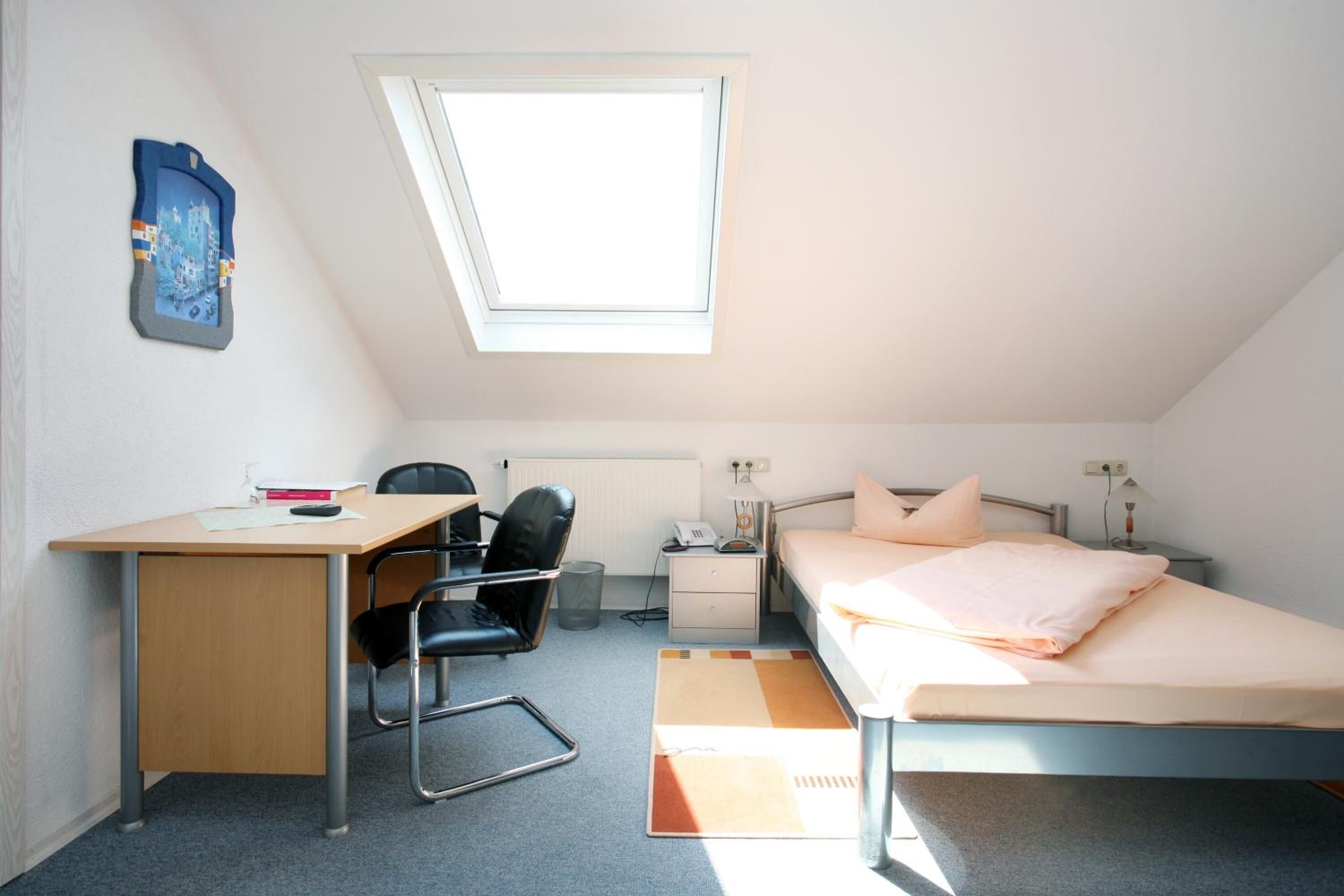 A small, well-lit room with a desk, chair, bed, and a window.