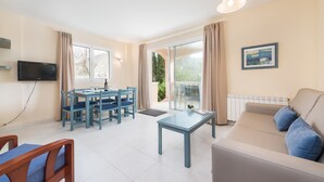 A modern living room with a sofa, a dining table with chairs, a TV, and a large window with curtains. - ORO DEL MAR IV Apartamento O2 - Apartment near the beach and with shared pool Free WiFi (Capdepera)