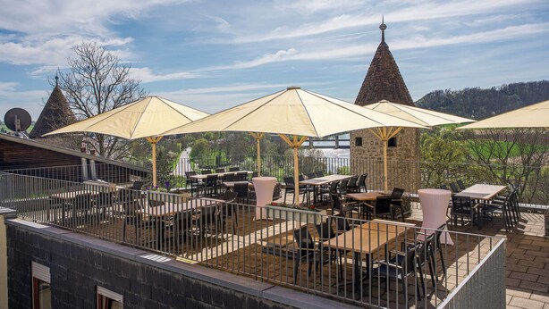 An outdoor patio with tables and chairs, umbrellas, and a view of a mountain with conical roofs. - Schloss Horneck (Gundelsheim)