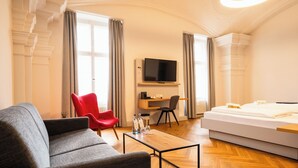 A hotel room with a bed, a grey sofa, a red chair, a wooden coffee table, a TV mounted on the wall, and a desk with a chair. - Schloss Horneck (Gundelsheim)