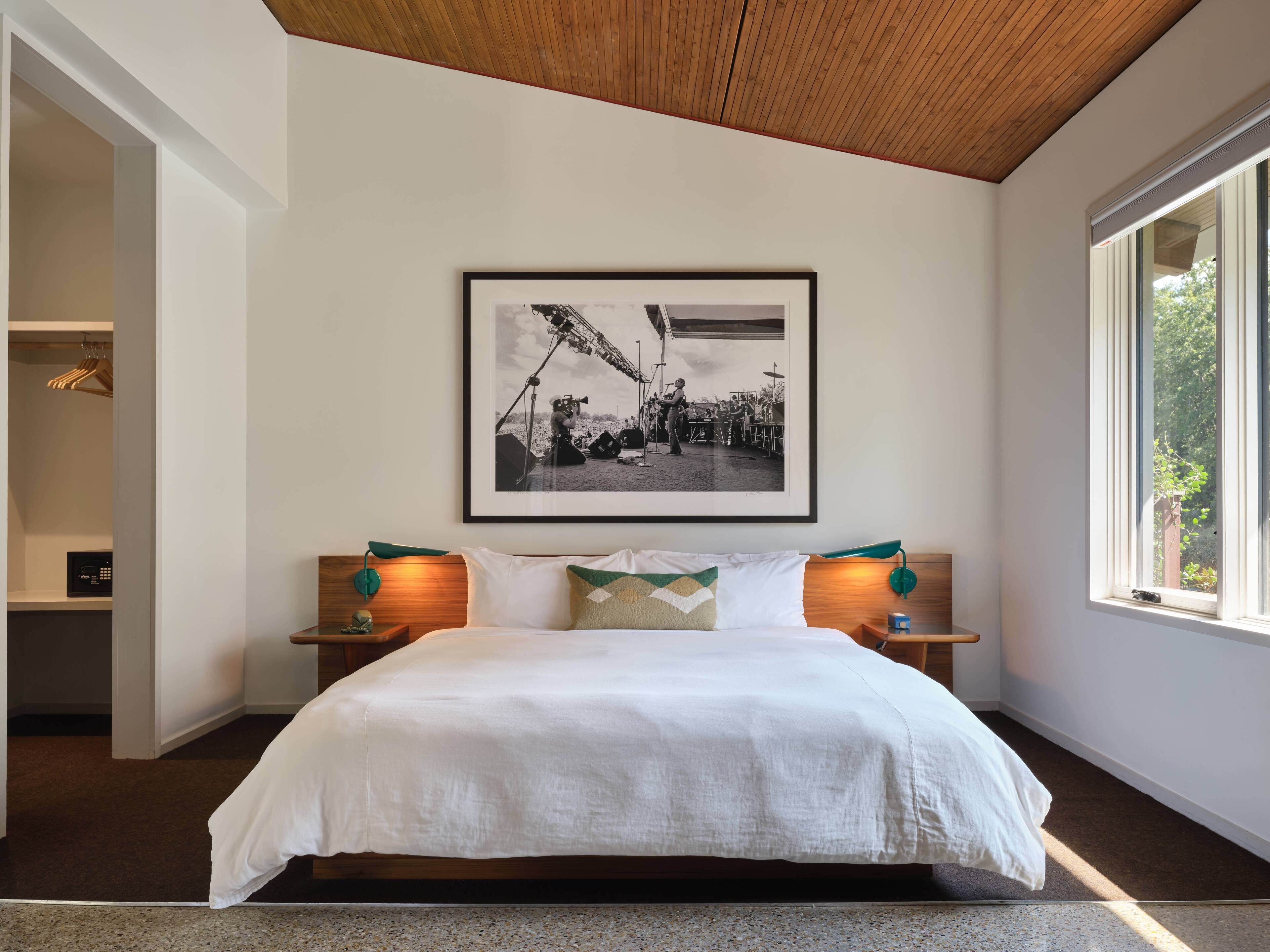 A bedroom with a large bed, a framed photograph on the wall, bedside lamps, and a window with a view of greenery.
