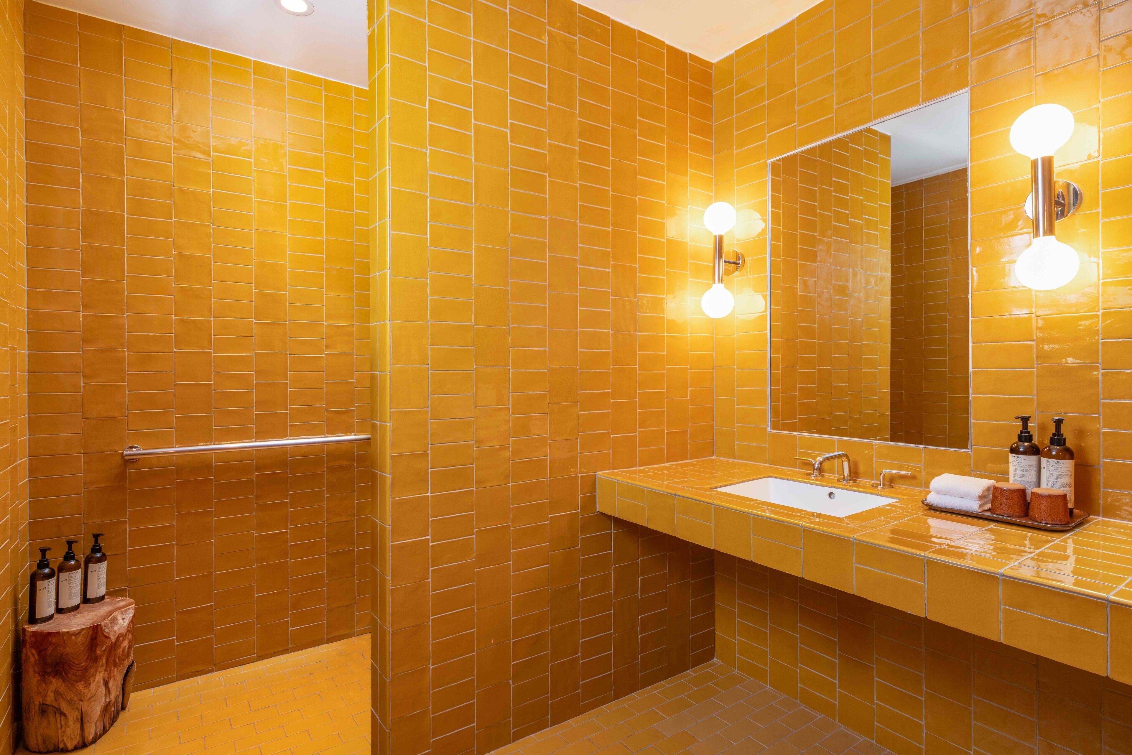 Une salle de bain avec des murs recouverts de carreaux jaunes, un lavabo blanc et un miroir avec des lumières.