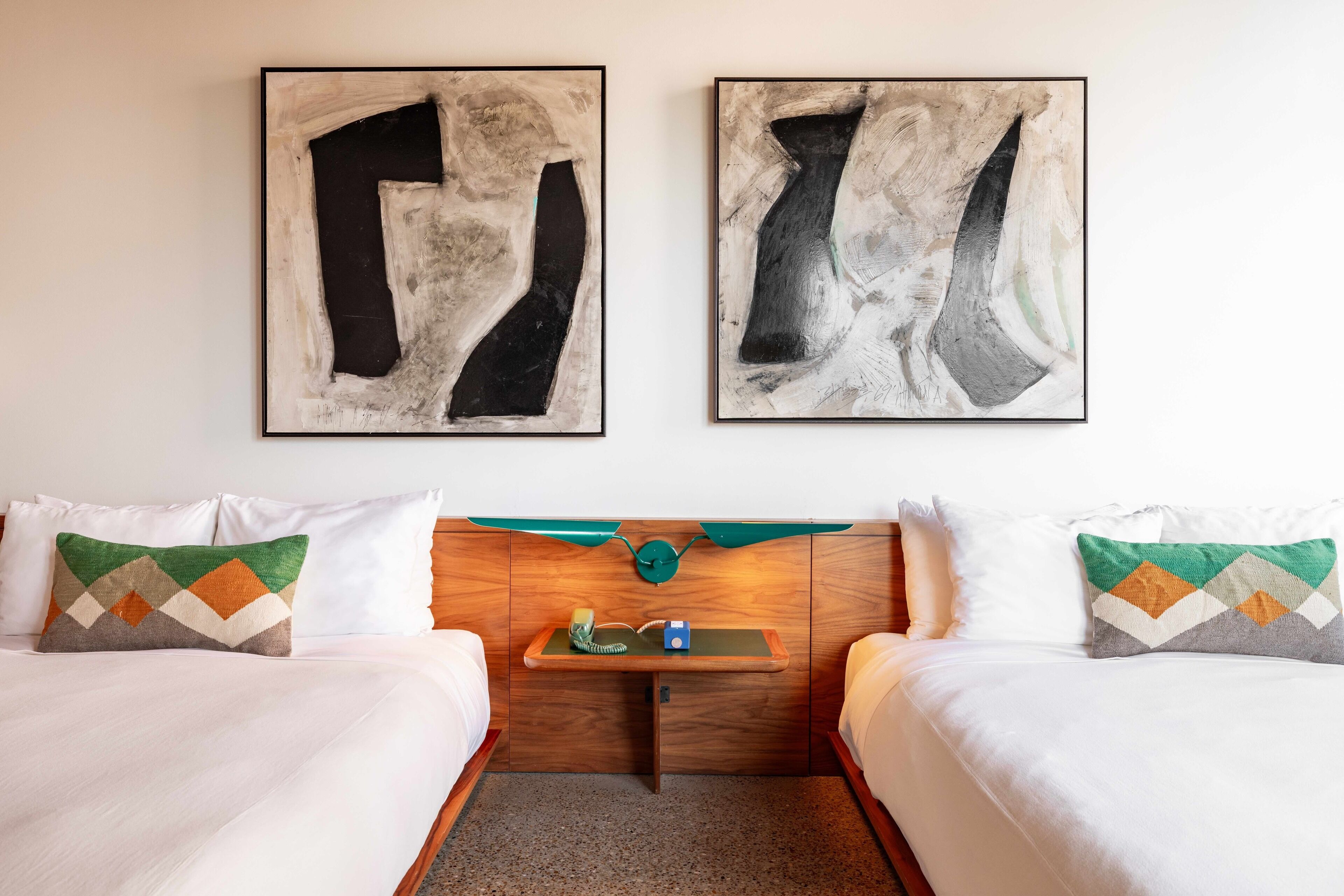 A hotel room with two beds, a wooden headboard, a small bedside table, and abstract artwork on the wall.
