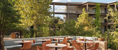Un espace extérieur aménagé en terrasse, avec des coins salon comprenant des canapés et des tables, entouré de verdure et avec un immeuble à plusieurs étages en arrière-plan.