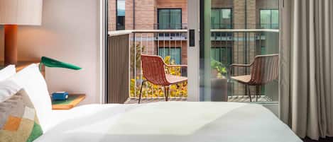 A hotel room with a large bed, a balcony with wicker chairs, and a view of a brick building.