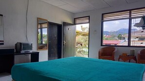 A room with a bed, a desk with a television, two chairs, and a window with a view of buildings and mountains. - OYO 2322 Dien Karona Hotel (Berastagi)