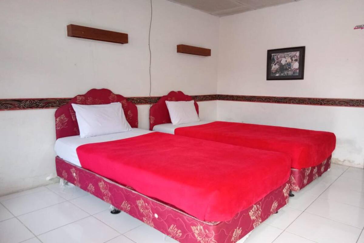 Quarto com duas camas, cada uma com colcha vermelha e travesseiros brancos. Possui duas cabeceiras de madeira e um quadro pendurado na parede.