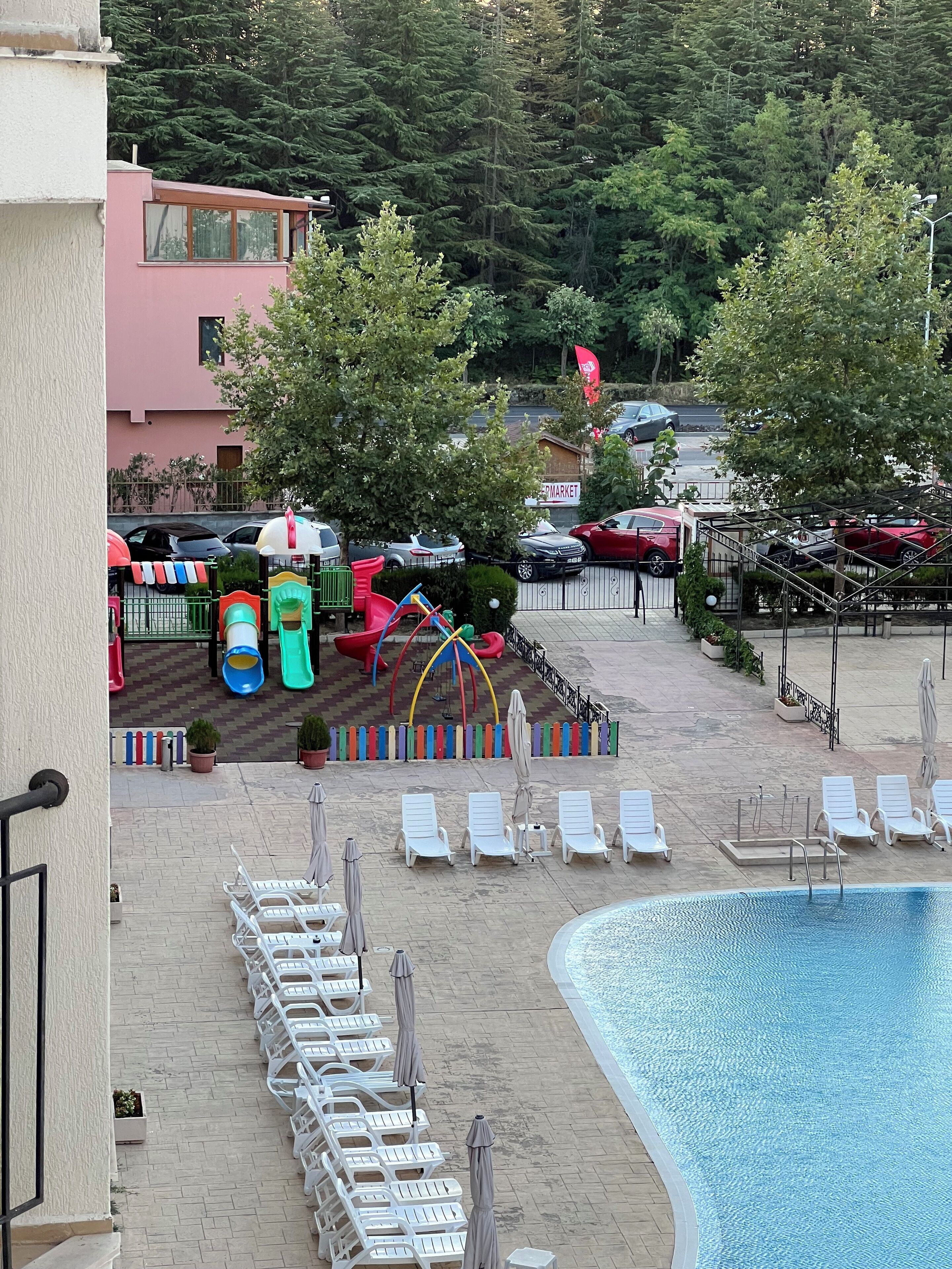 A pool area with lounge chairs and umbrellas, a children's play area with slides, and a parking lot with cars.