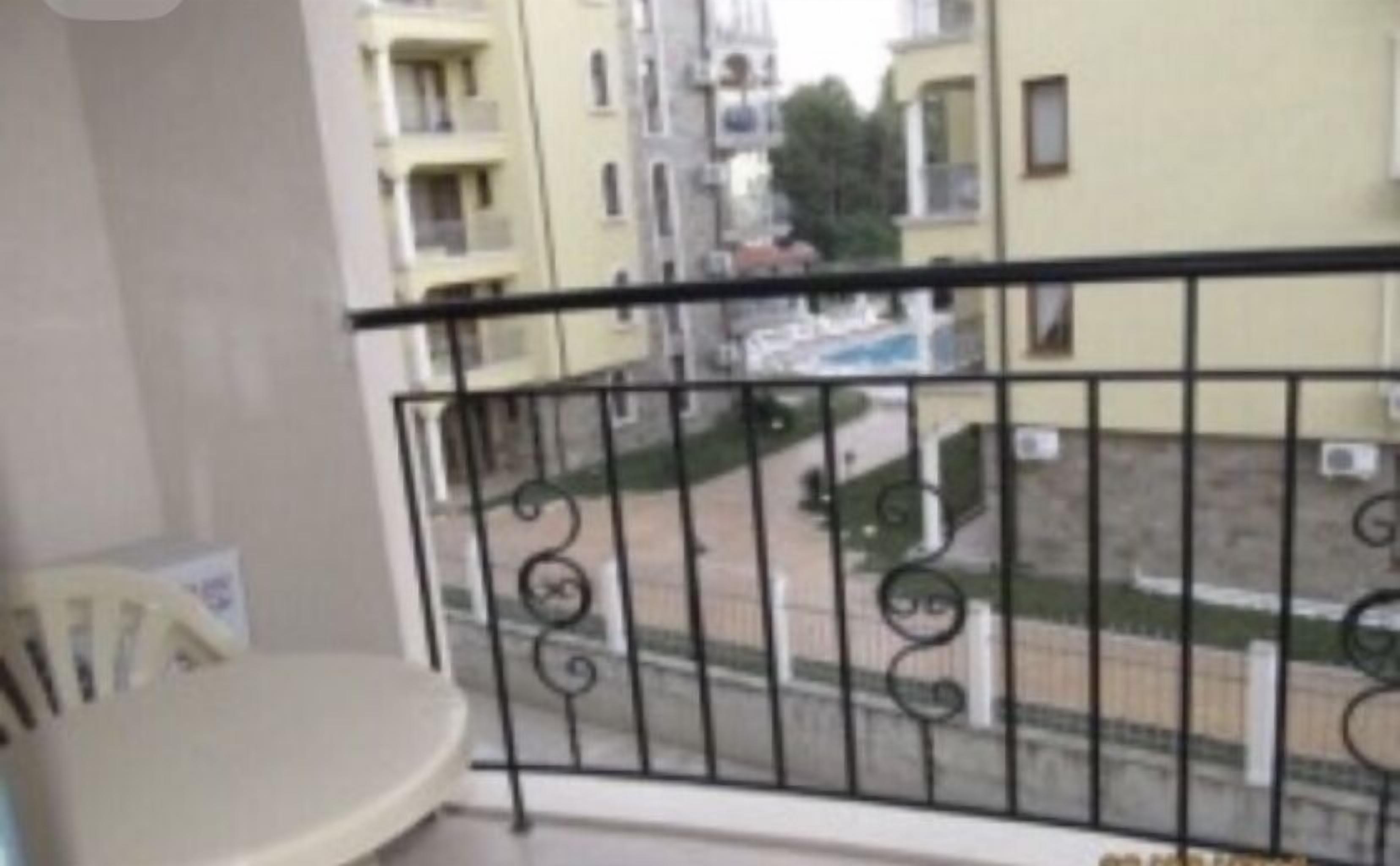 Un balcó amb barana metàl·lica, una cadira blanca i vista a una zona residencial amb edificis i una piscina.