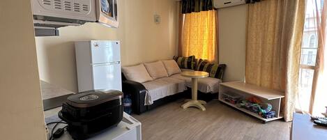 A compact living space with a kitchenette, a sofa, a small table, and a window with curtains.