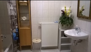 Une salle de bain avec un lavabo blanc, un miroir, un radiateur, un petit tabouret et une étagère contenant des articles de toilette.