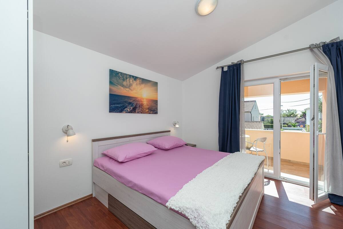 A bedroom with a bed, a painting, a window, and a view of the outside.