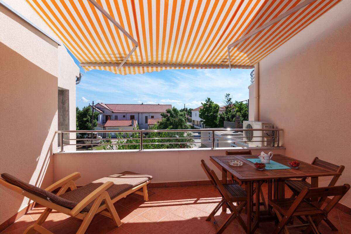 A balcony with a striped awning, wooden furniture, and a view of buildings in the background.