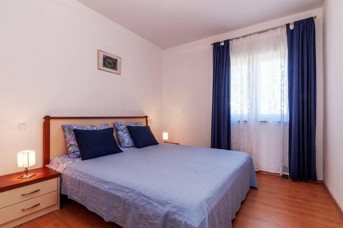 A neatly made bed with a blue bedspread and pillows, a bedside table with a lamp, and a window with blue curtains.