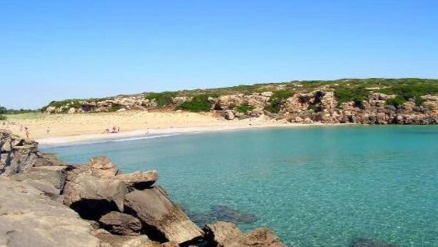 A coastal beach with clear turquoise water, rocky formations, and a sandy shore.