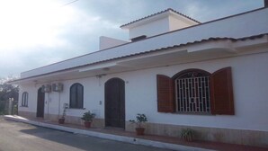 A single-story building with a flat roof, white exterior walls, and a small attached garage. There are potted plants along the front walkway, and the building has a simple, functional design with minimal decorative elements.