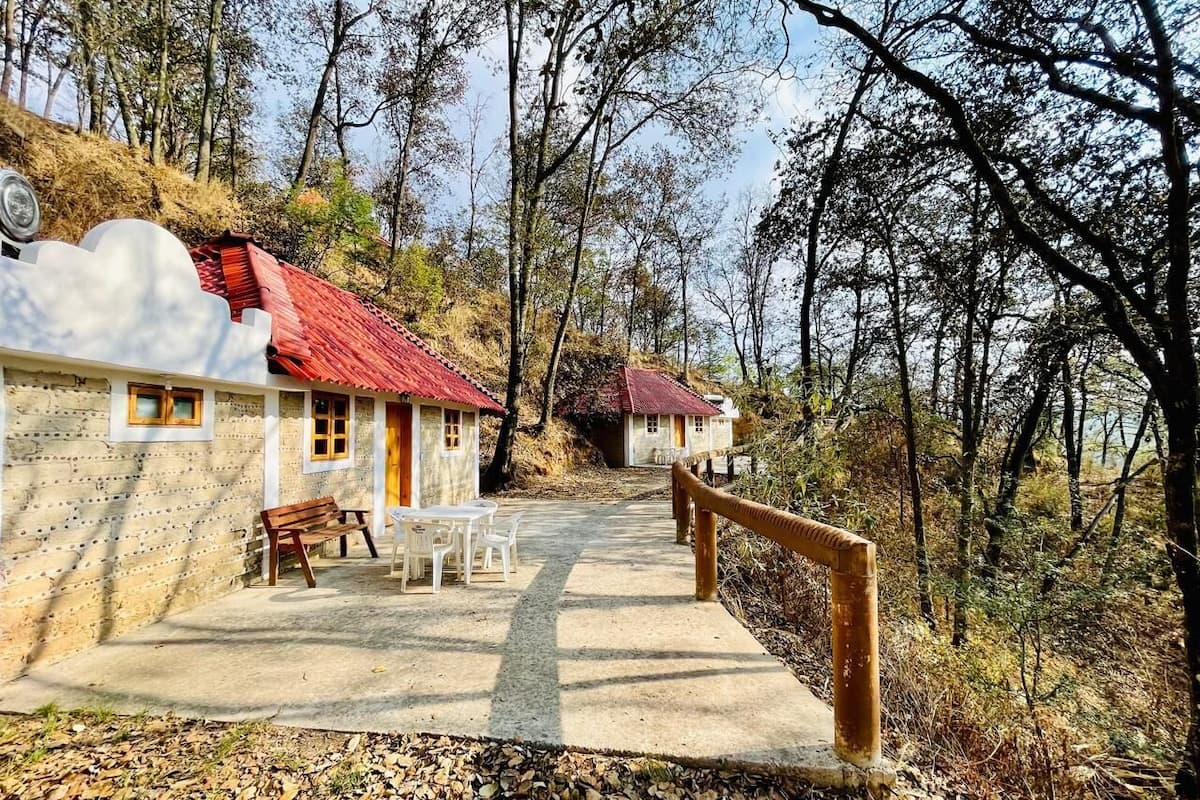 A rustic building with a red roof, a concrete pathway leading to a wooden bench and table, surrounded by trees.