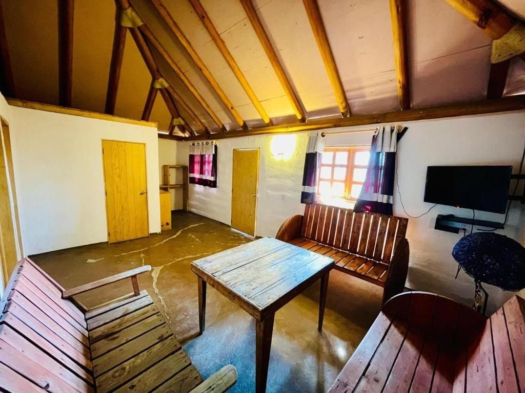 Una habitación con una mesa de madera, un banco, un televisor y una ventana con cortinas.