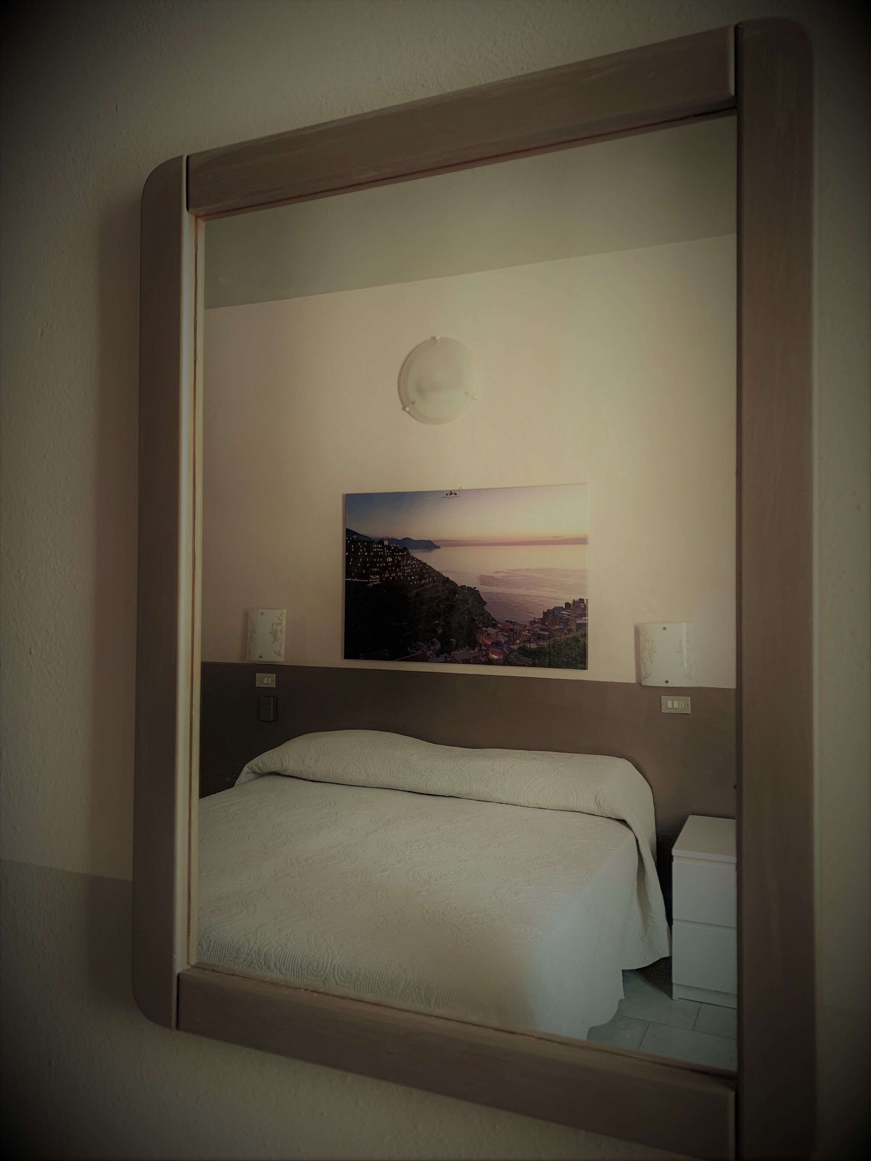 Une chambre d’hôtel avec un lit, une table de chevet, un luminaire fixé au mur et un tableau représentant un paysage côtier.