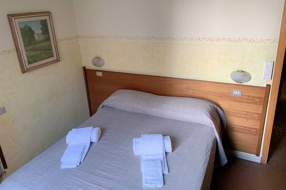 A neatly made bed with two towels, a wooden headboard, a wall-mounted light fixture, and a framed picture.