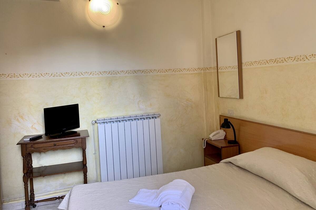 A hotel room with a bed, a desk with a computer, a radiator, a telephone, and a mirror on the wall.