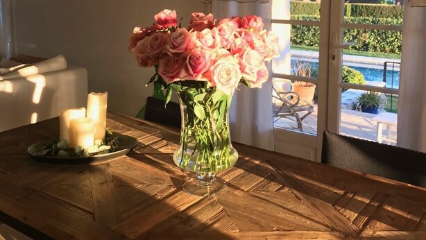 Une table en bois ornée d’un vase de roses roses, d’une bougie allumée et offrant une vue sur un jardin par la fenêtre.