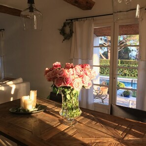Une table en bois ornée d’un vase de roses roses, d’une bougie allumée et offrant une vue sur un jardin par la fenêtre.