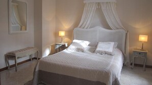 Quarto com cama de dossel, mesas de cabeceira com luminárias, um banquinho e um espelho.