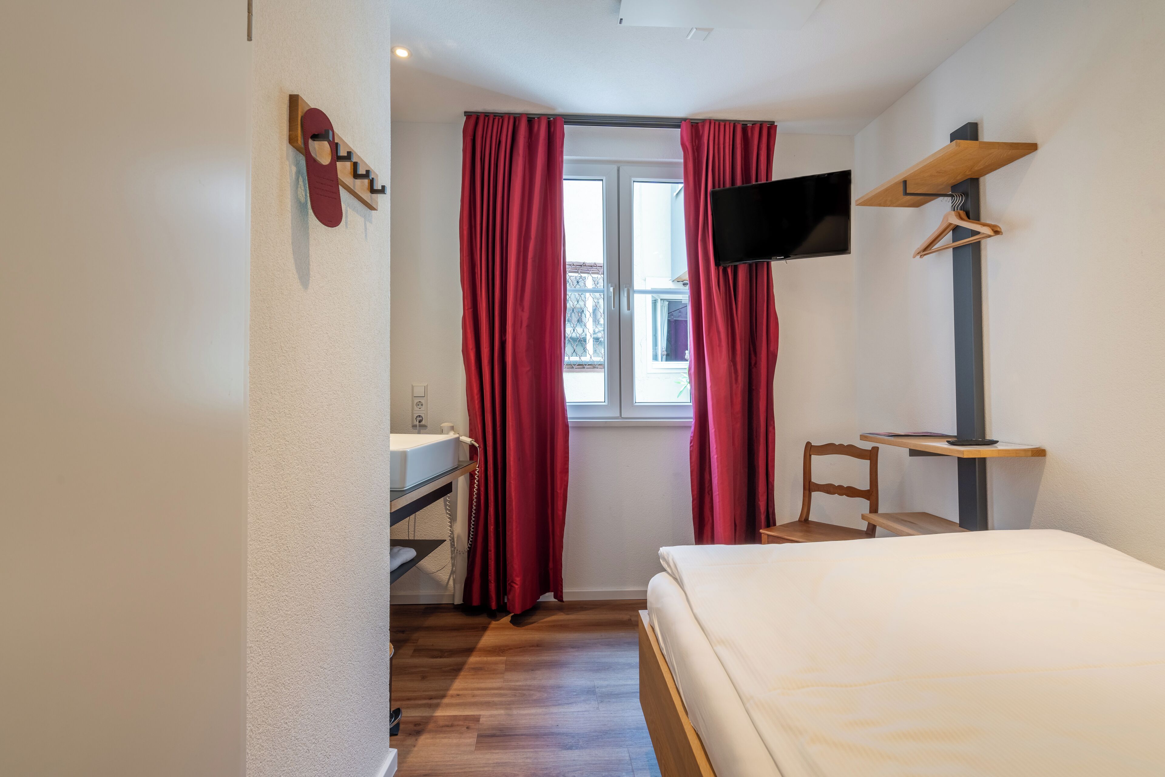 Une chambre d’hôtel comprenant un lit, un bureau avec une chaise, un téléviseur fixé au mur et une fenêtre avec des rideaux.