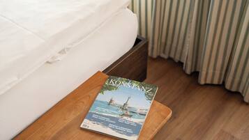 Ein Bett mit weißen Bettwäsche, ein Nachttisch aus Holz mit einer Zeitschrift mit dem Titel 'KONSTANZ' darauf und gestreifte Vorhänge im Hintergrund.
