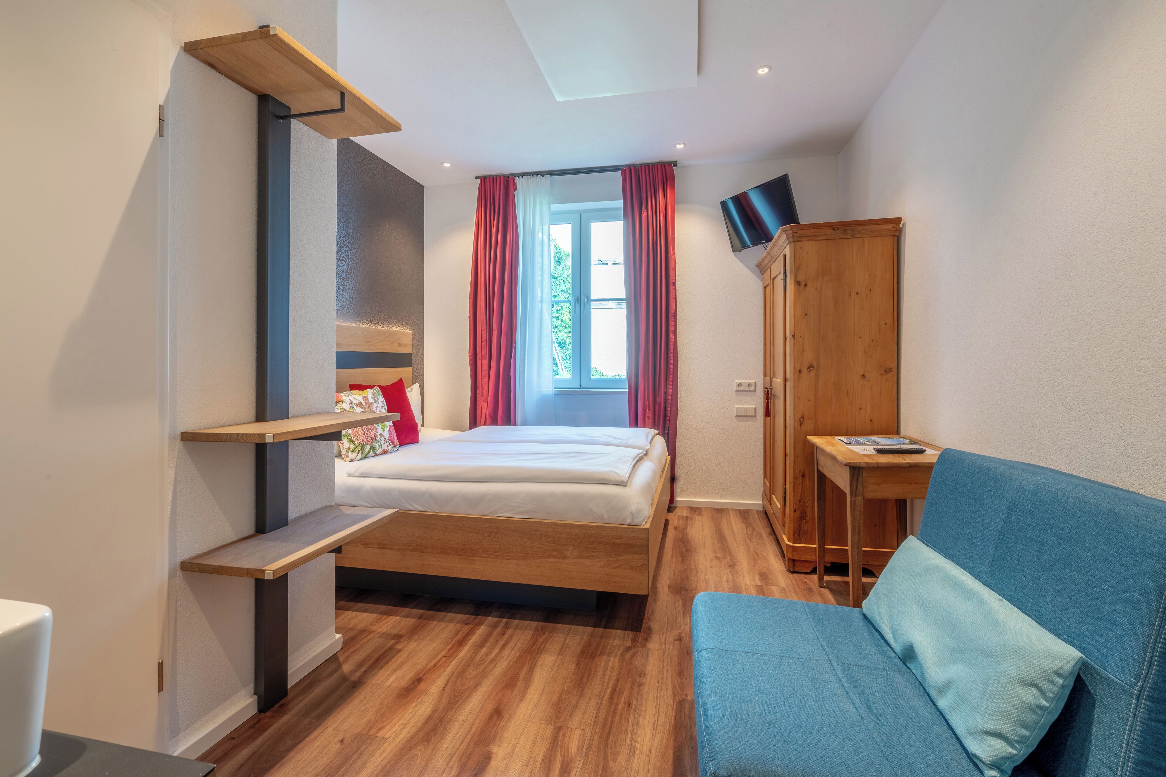 Une chambre à coucher avec un lit, un canapé bleu, une armoire en bois, un téléviseur fixé au mur et une fenêtre avec des rideaux.