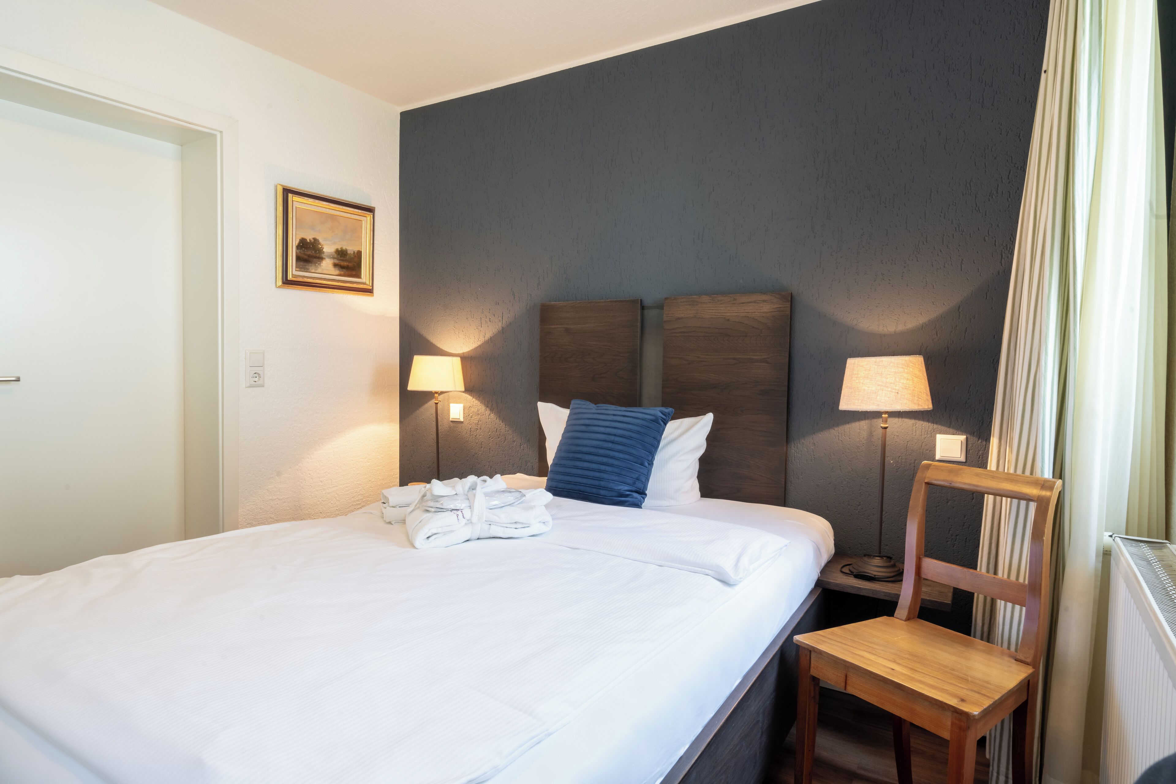 A neatly made bed with white linens, two bedside lamps, a wooden chair, and a framed picture on the wall.