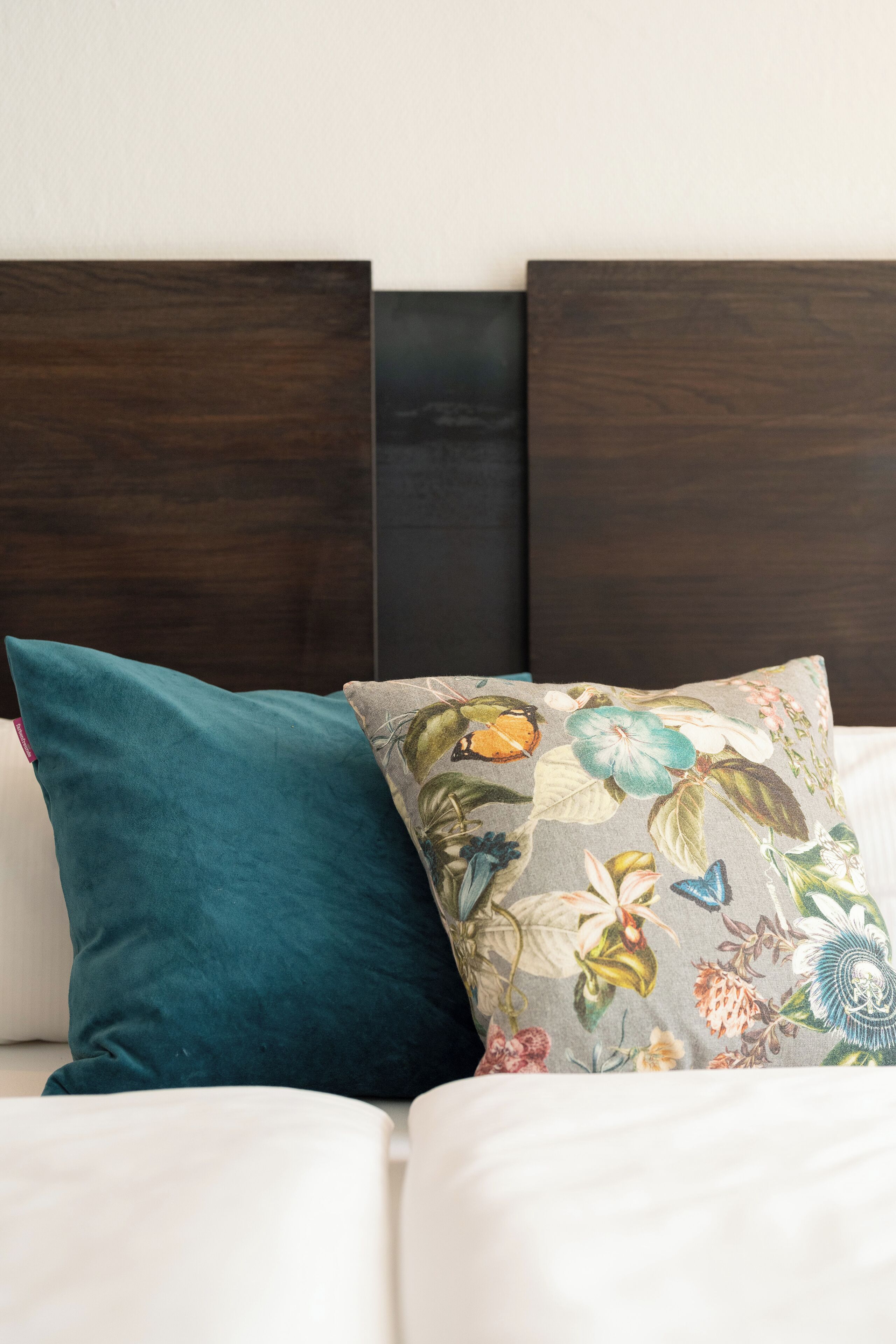 Una cama con cabecera oscura y dos almohadas, una de color turquesa liso y otra con estampado floral.