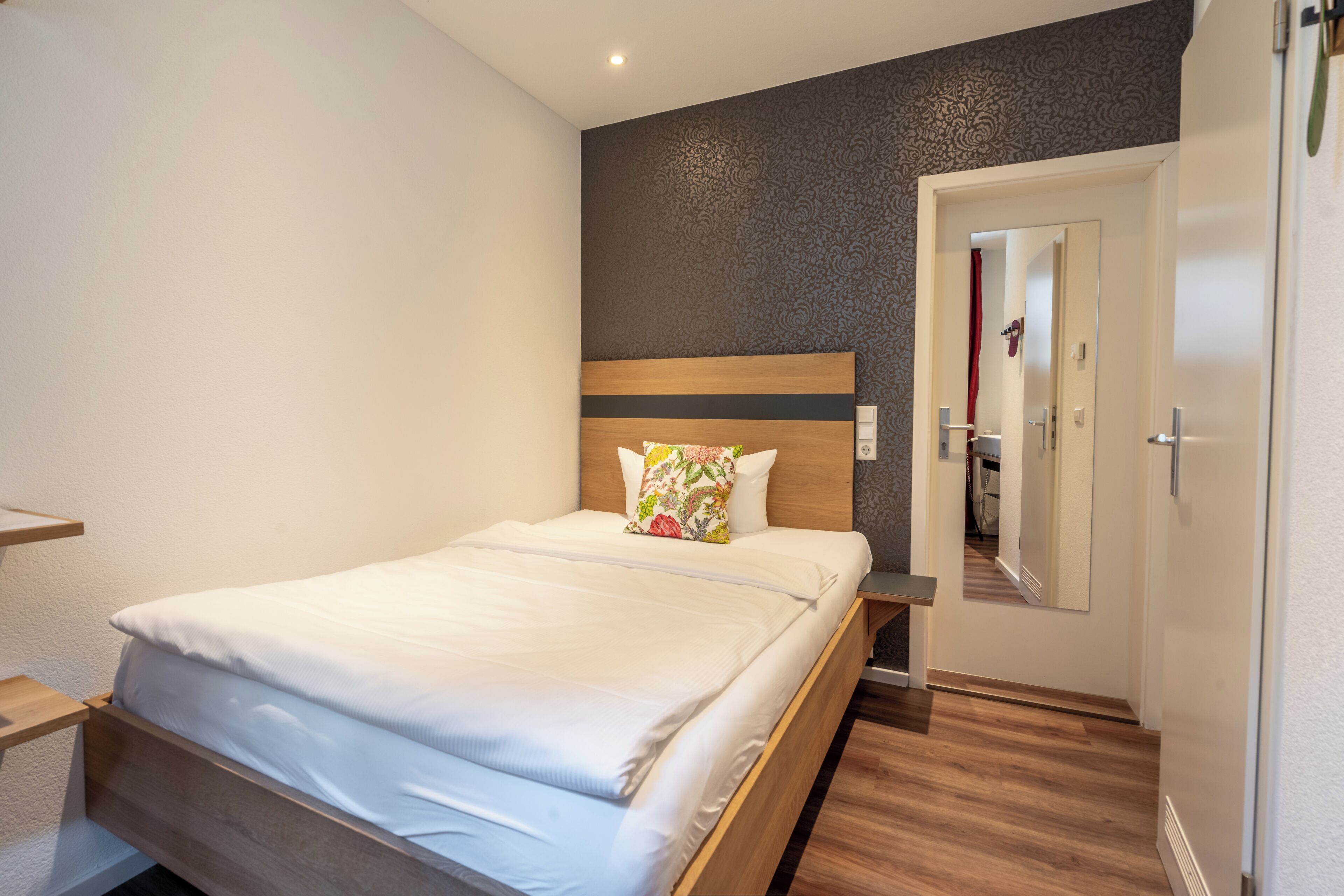 A modern hotel room with a wooden bed, a nightstand, a mirror, and a door leading to a bathroom.