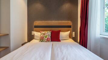 Una cama bien hecha con sábanas blancas y dos almohadas, una con motivos florales y otra roja, en una habitación con pared estampada y una ventana con cortinas rojas.