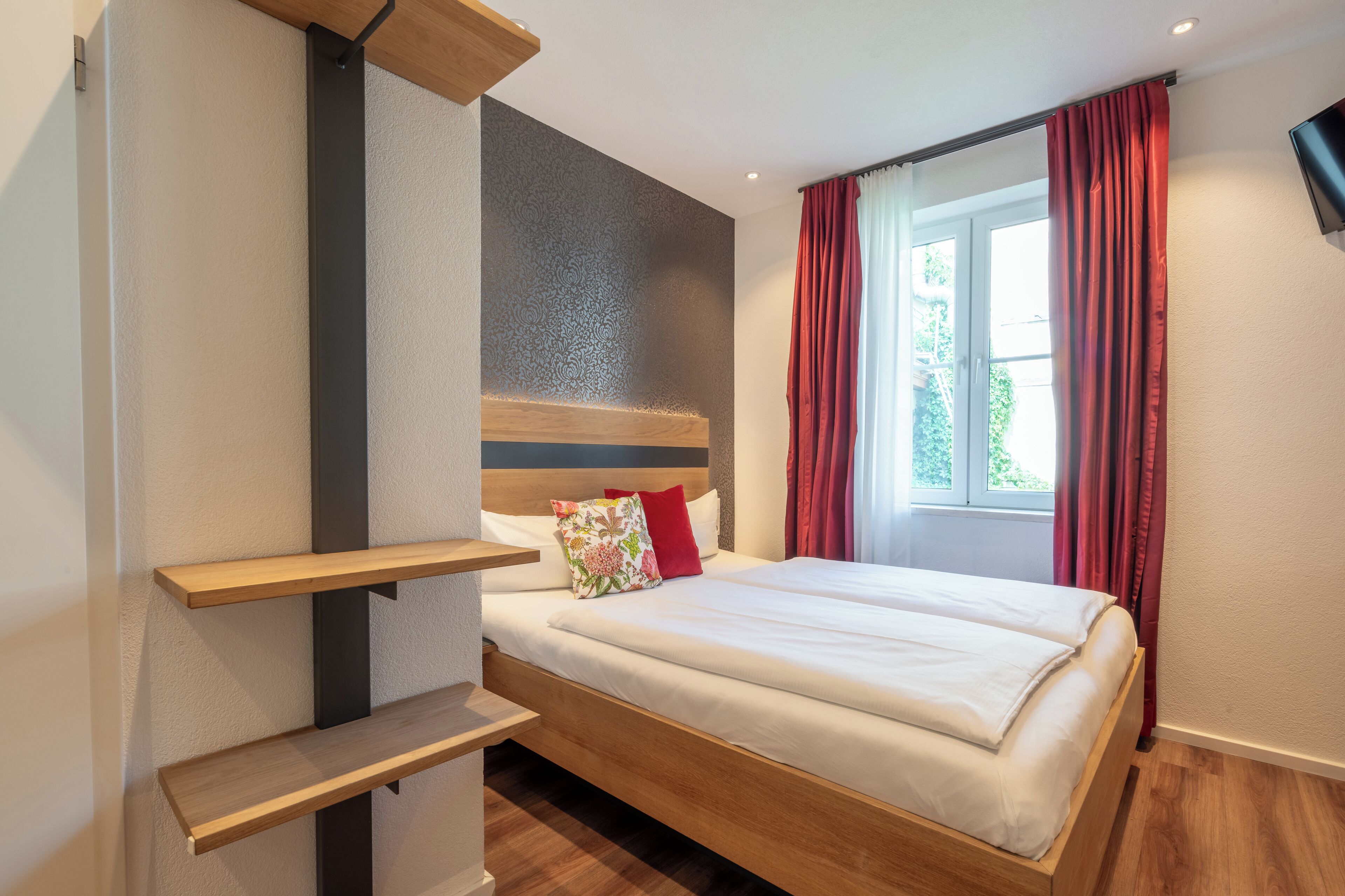 Een moderne hotelkamer met een houten bed, een klein houten plankje en een groot raam met rode gordijnen.
