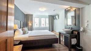 A modern hotel room with a large bed, a wooden floor, a bedside table with a lamp, a bathroom with a sink and mirror, and a view of the outside through the window. - Hotel am Fischmarkt (Konstanz)
