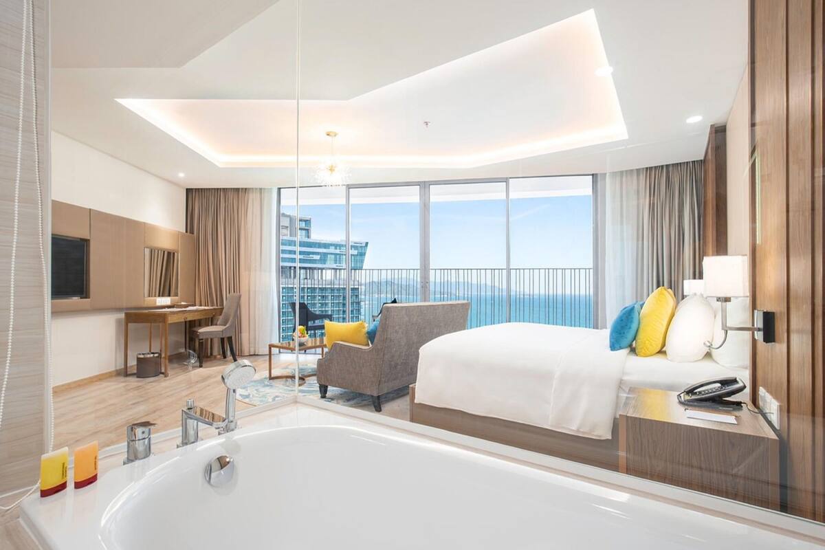 A modern hotel room with a large bed, a sitting area with a chair and table, a bathtub, and a large window offering a view of the city.
