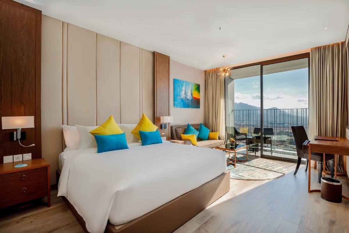A hotel room with a large bed, a desk, and a balcony with a city view.