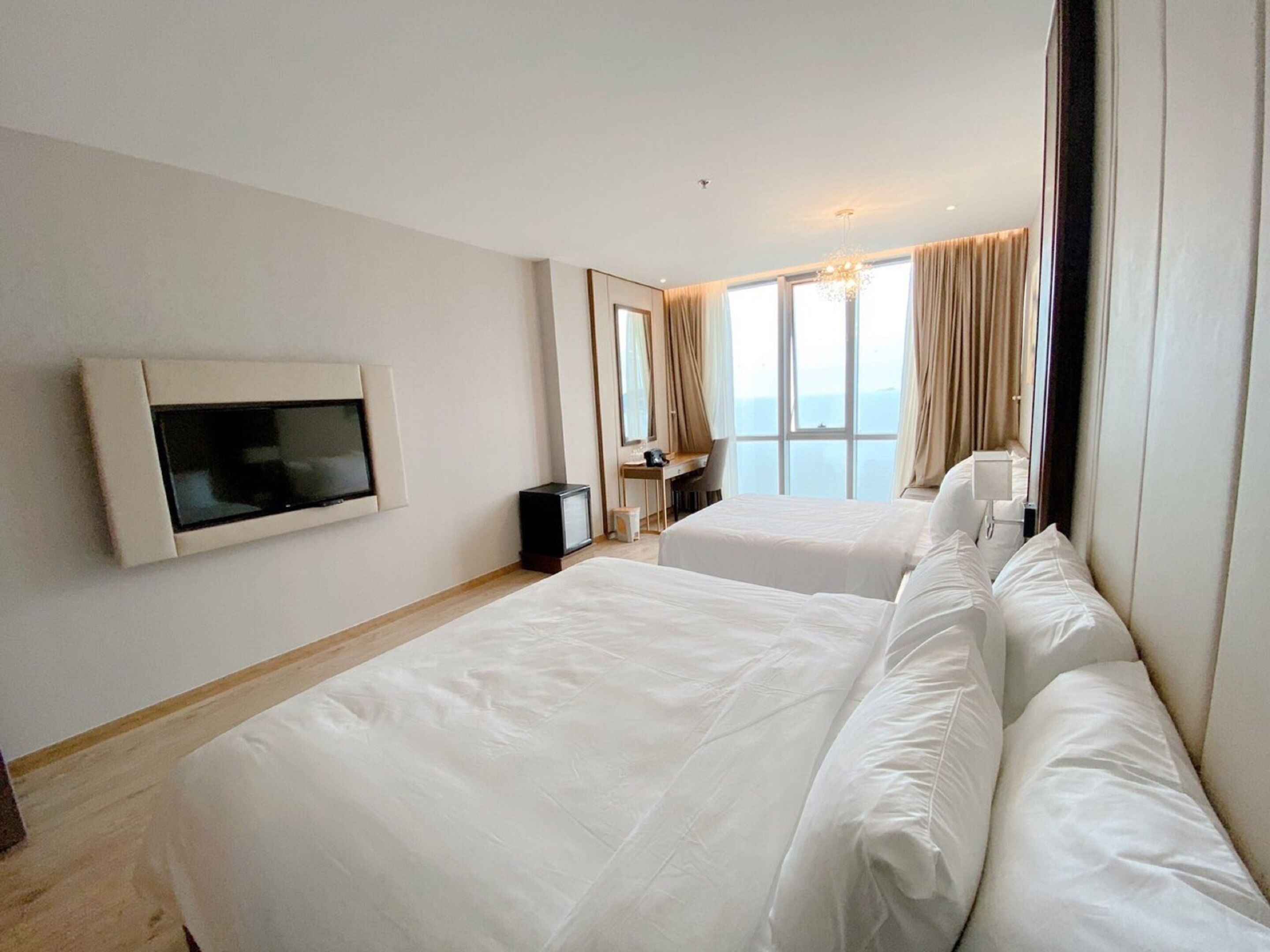 A hotel room with two beds, a TV mounted on the wall, a desk with a chair, and a large window with curtains.