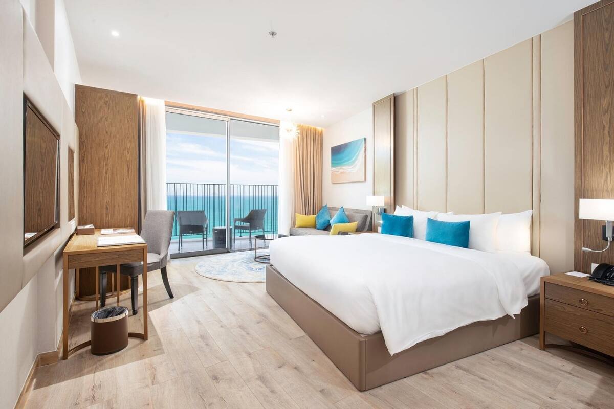 A hotel room with a large bed, a desk, a chair, a small table, a trash bin, a nightstand, and a view of the ocean.