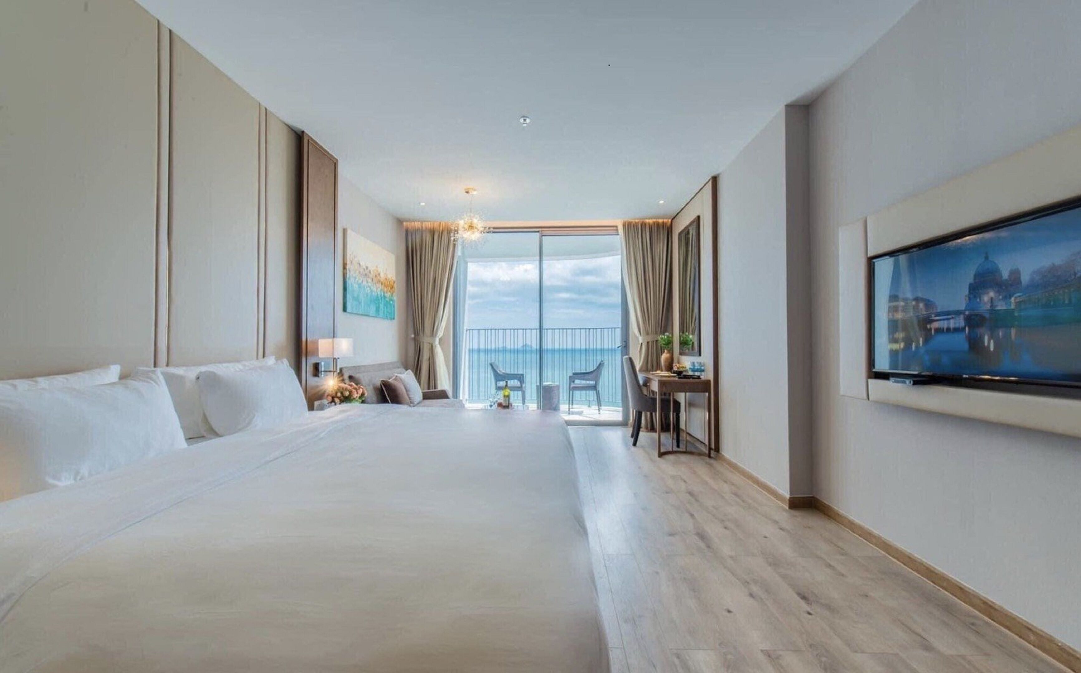 A hotel room with a large bed, a desk with two chairs, a TV, and a balcony with a view of the sea.