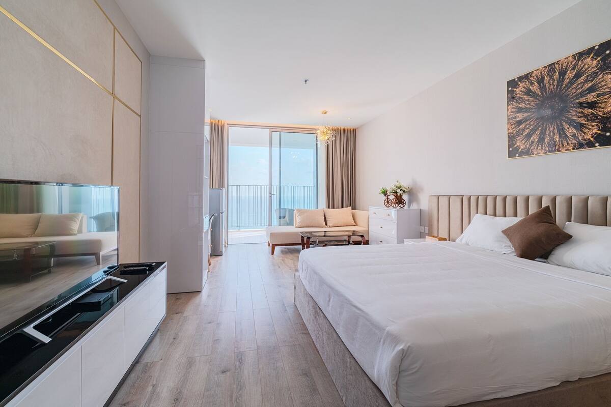 A modern hotel room with a large bed, a sofa, a TV, and a balcony with curtains.