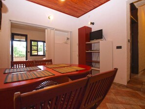 A dining area with a wooden table and chairs, a television on a shelf, and a window with a view of greenery.