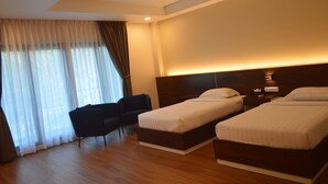 Habitación de hotel con dos camas individuales, un escritorio, una silla y un ventanal con cortinas.