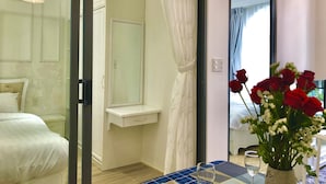 Ein Hotelzimmer mit einem Bett, einem Nachttisch, einem Spiegel, einem kleinen Schreibtisch mit Stuhl, einer Vase mit Blumen und einem Fenster mit Vorhängen.